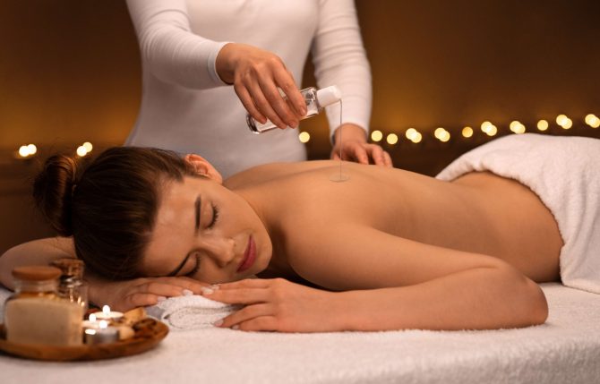 Unrecognizable massage therapist pouring oil on sleeping lady back at luxury spa decorated with candles, body care concept. Young woman enjoying relaxing massage with aromatherapy